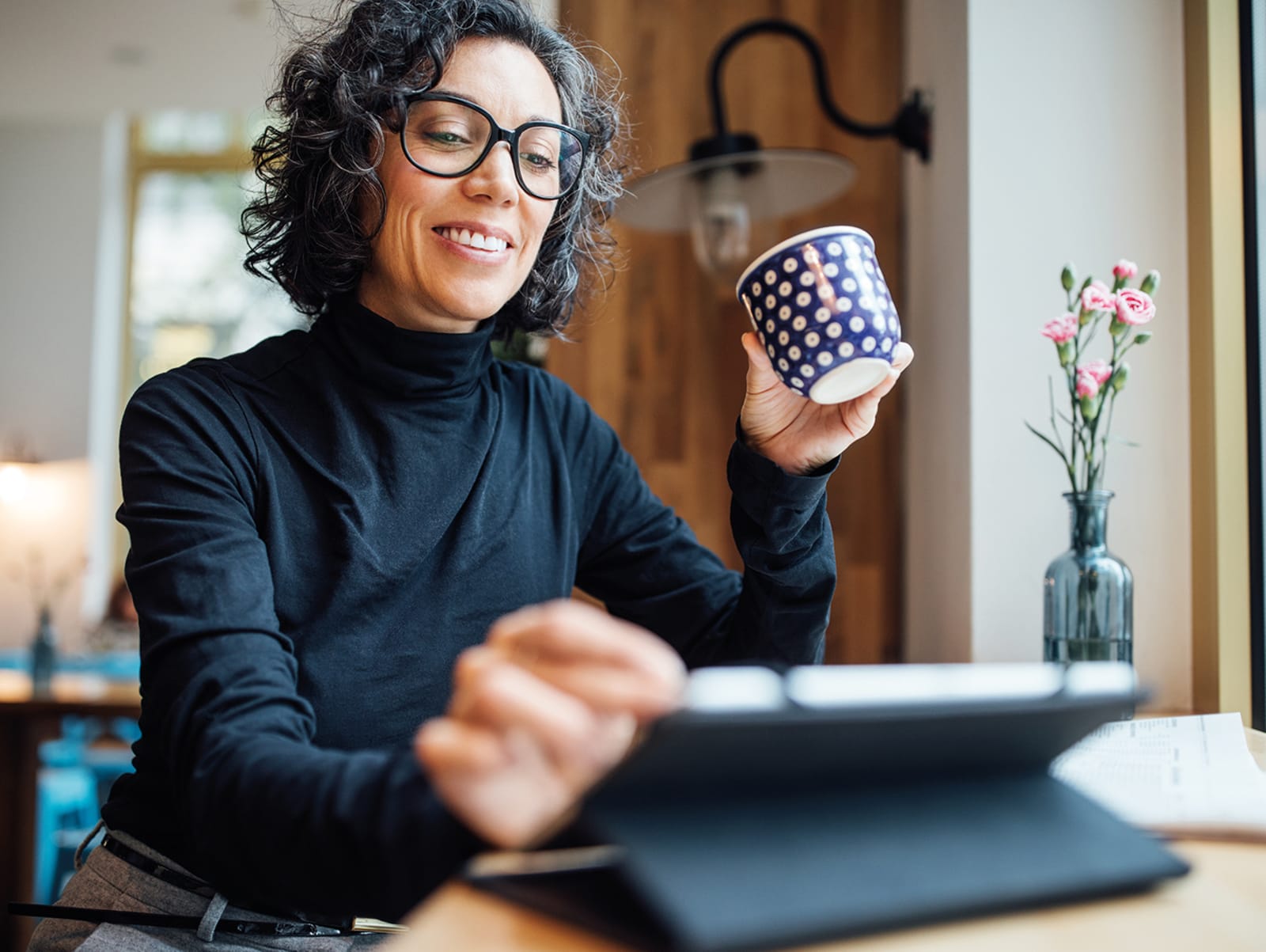 Woman wearing black roll neck smiling at tablet whilst drinking a coffee from a navy and white spotted mug