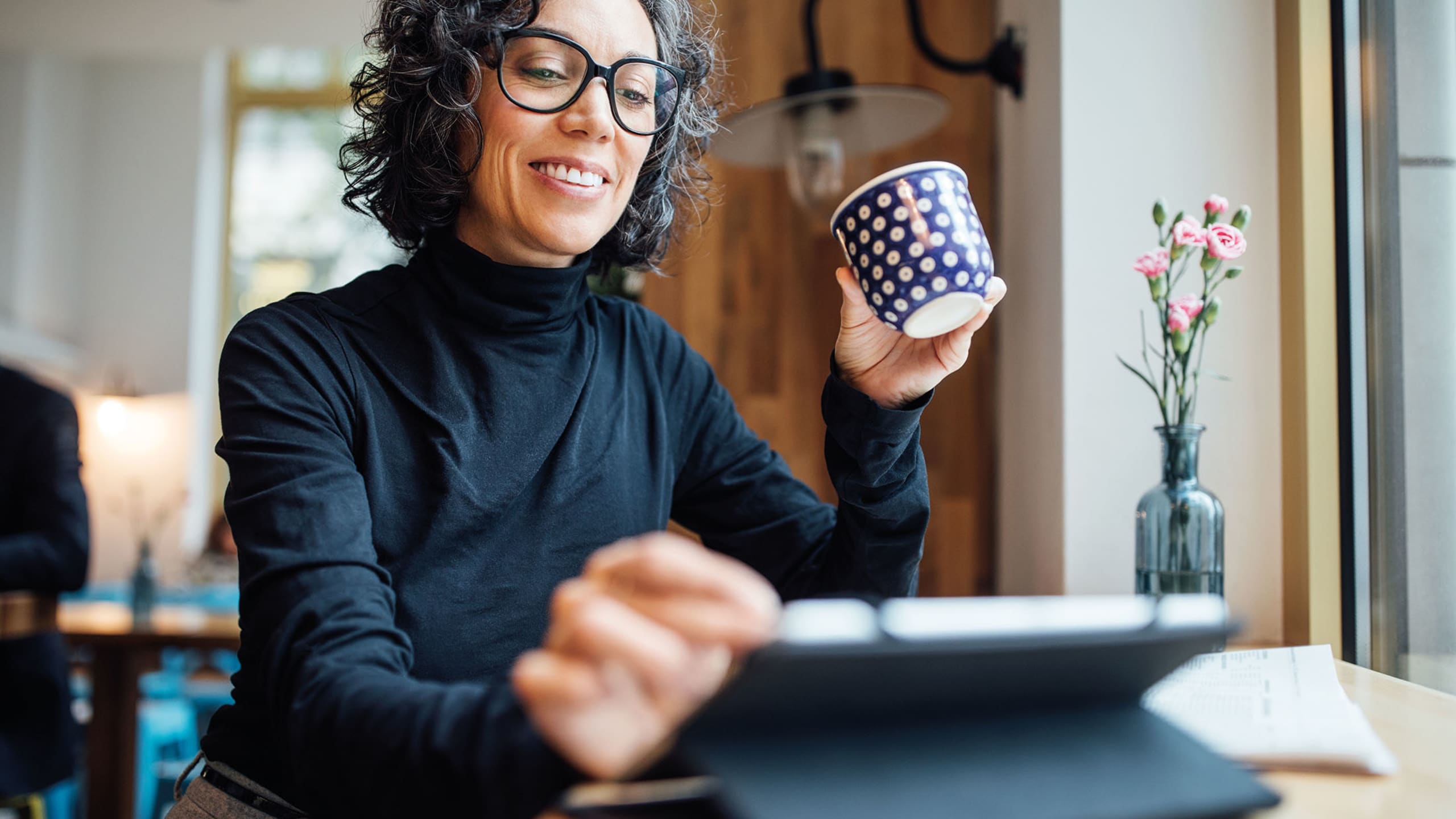 Woman wearing black roll neck smiling at tablet whilst drinking a coffee from a navy and white spotted mug